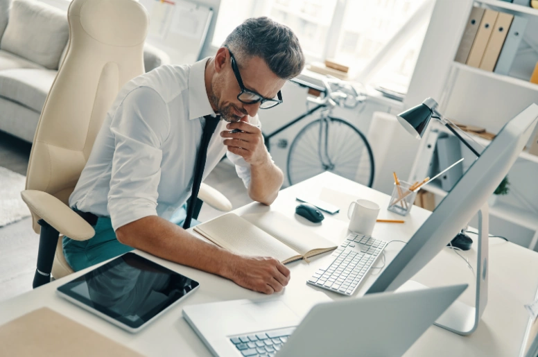 A man in a shirt and tie sits at a desk, working on a laptop and tablet, focused on his SEO tasks.