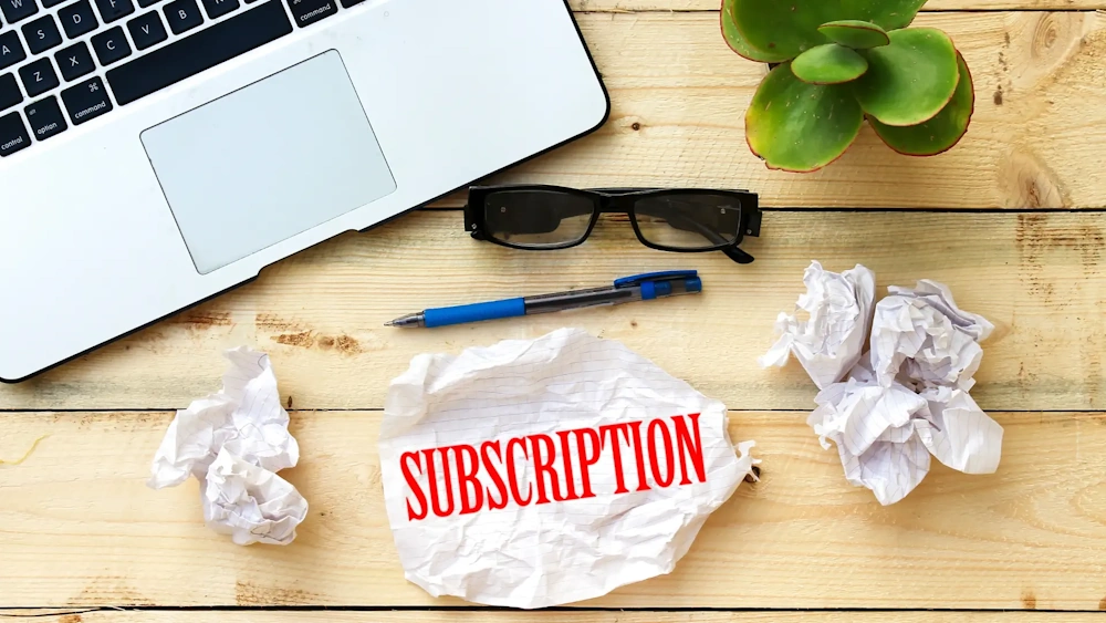 Wooden table with a laptop, glasses, pen, succulent plant and one paper shows "Subscription" indicating subscription services
