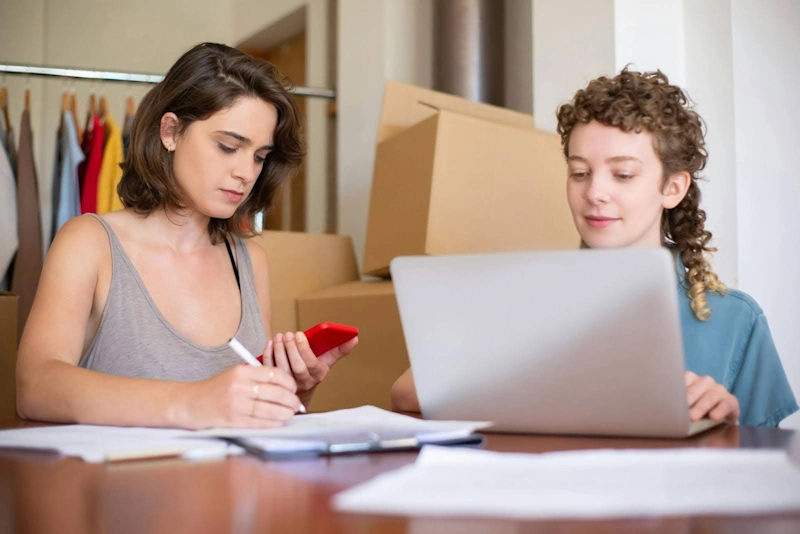 Email marketing for small businesses teamwork Two women working at a desk with a laptop, smartphone, and paperwork, focusing on email marketing for small businesses.